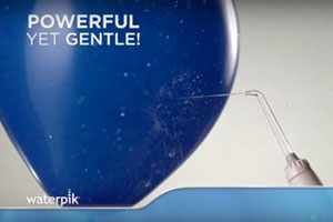 Blue balloon with the words Powerful Yet Gentle! with a Waterpik™ water flosser handle and tip shooting water against it