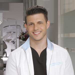 Photo of Dr. Chris Strandburg, indoors in his dental office, with short brown hair, wearing a white lab coat and black shirt