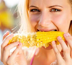 Image of a blonde woman, outside, wearing a pink tank top, eating corn on the cob