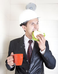 Image of a brunette man in shower with water on, wearing suit, tie, and hard hat, holding a red coffee cup, eating a sandwich