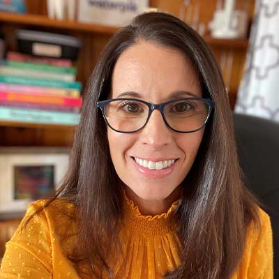 Photo of Sue Scherer smiling. She has long brown hair, large framed glasses, and an orange sweater. Behind her is a bookcase with books on their side.